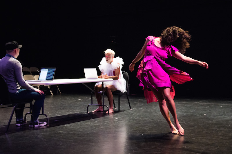 Michele Boule, in a fuschia dress, dances in front of two men seated at narrow table with computers in front of them